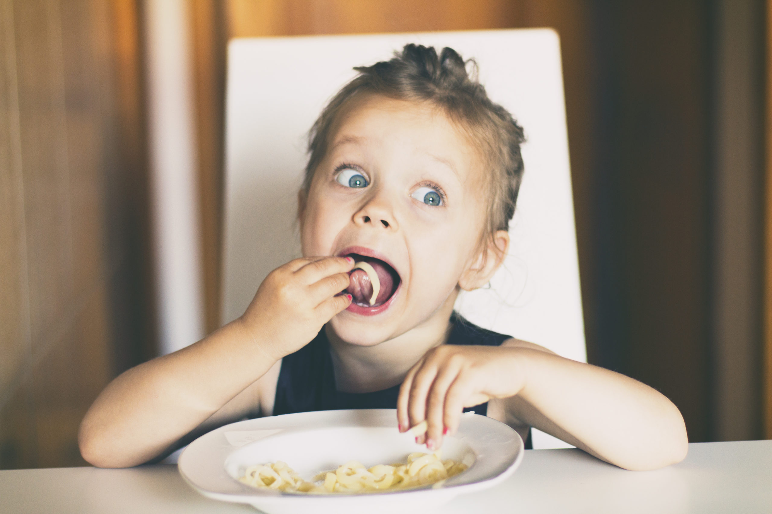 Three years old girl sitting at the table in the kitchen and eating pasta.